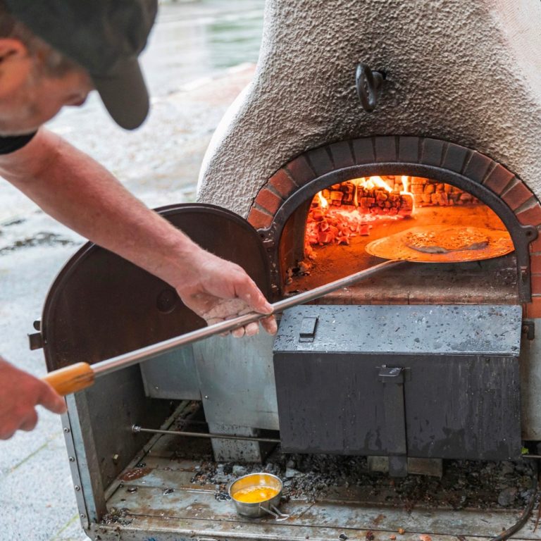 Eine Person schiebt mit einer langen Pizzaschaufel eine Pizza in einen großen, mit Holz befeuerten Steinofen.