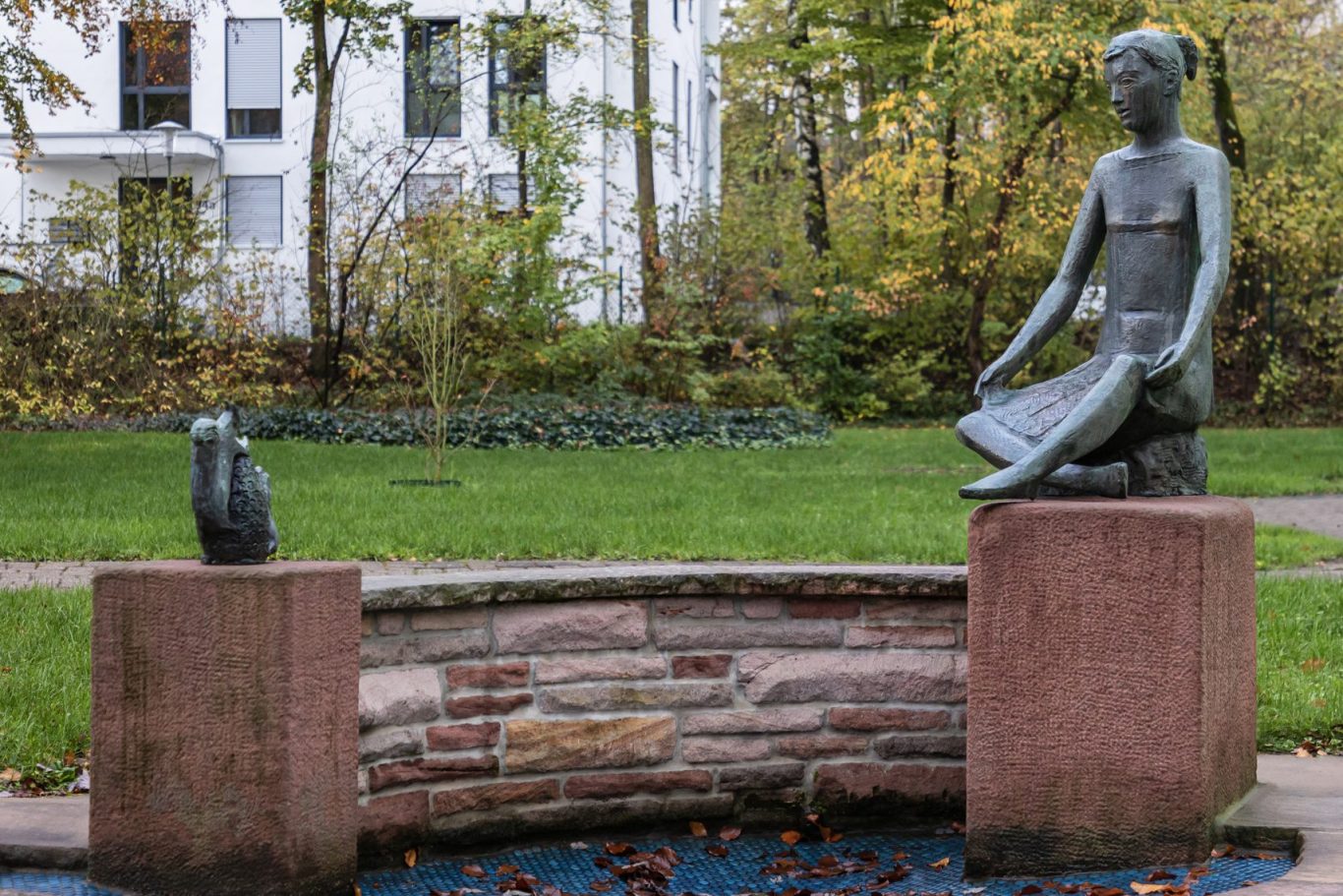 Bronzeskulptur einer sitzenden Frau auf rotem Sockel, gegenüber einer kleinen Tierfigur in einem begrünten Innenhof mit Bäumen und einer Ste