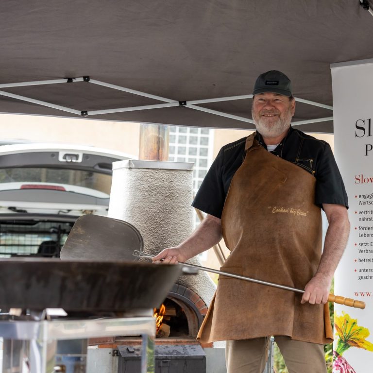 Mann mit Lederschürze bedient einen mobilen Holzbackofen unter einem Pavillon.