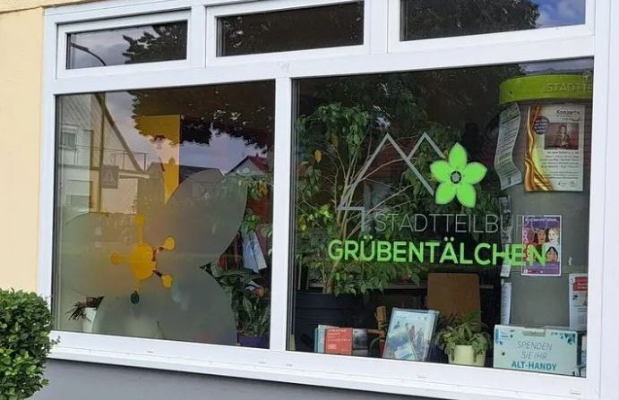Auf dem Foto ist das Fenster des Stadtteilbüros zu sehen, auf dem das Logo mit stilisierten Häusern und einer grünen Blüte abgebildet ist.