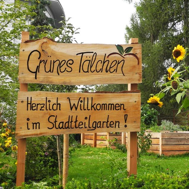Holzschild mit der Aufschrift „Grünes Täuchen – Herzlich Willkommen im Stadtteilgarten!“ in einem bepflanzten Garten mit Sonnenblumen.