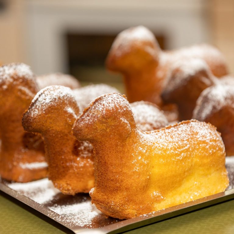 Mehrere goldbraune, mit Puderzucker bestäubte Gebäckstücke in Form kleiner Lämmer auf einem Backblech.