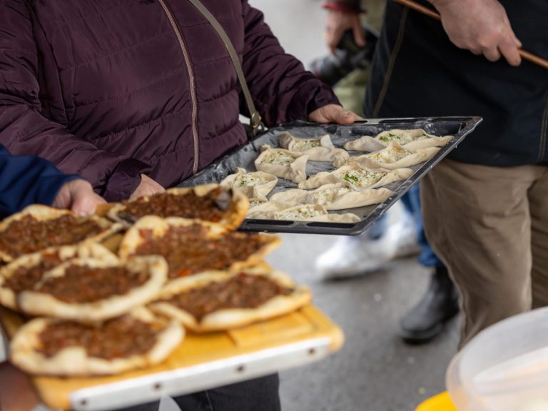 Menschen präsentieren auf Blechen frisch zubereitete Teigtaschen mit Käsefüllung und auf einem Brett kleine Fladenbrote mit würzigem Hackfle